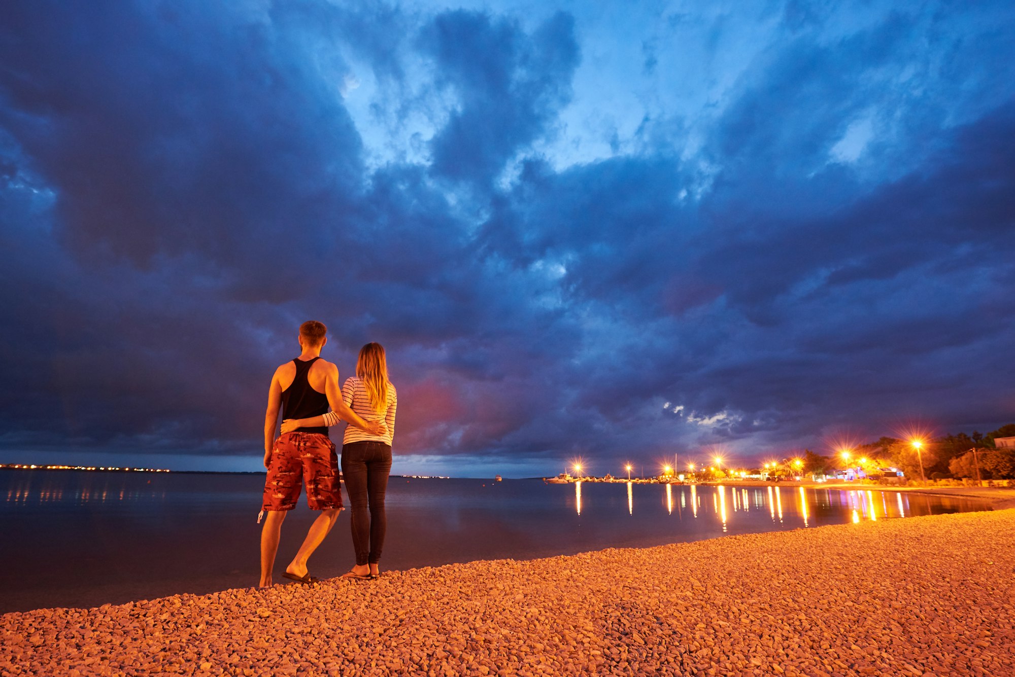 Man and woman resting on pebble beach at dusk on calm water dramatic blue cloudy sky background