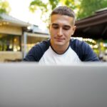 Young man sitting at table and typing on laptop keyboard while working in outdoor cafe