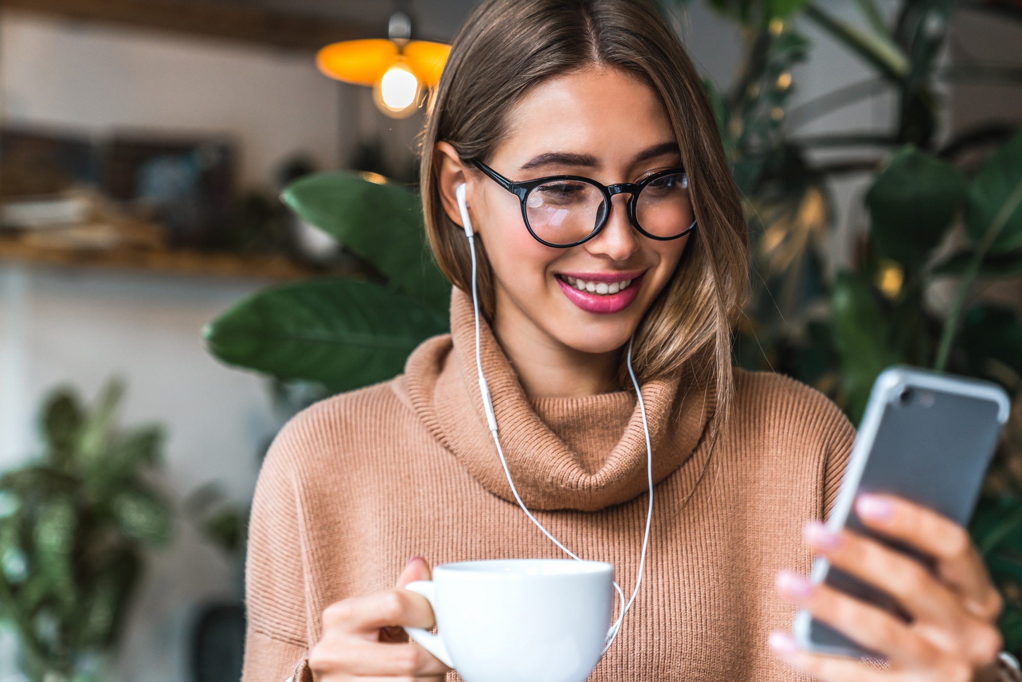 Woman sitting at a table with laptop and looking at mobile phone while drinking coffee